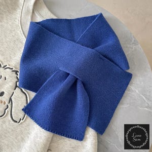 May include: A solid royal blue knit scarf with a simple rectangular shape and finished edges, layered over a light heather gray sweatshirt featuring a partial embroidered graphic of a dog with dark outlines and small brown accents.