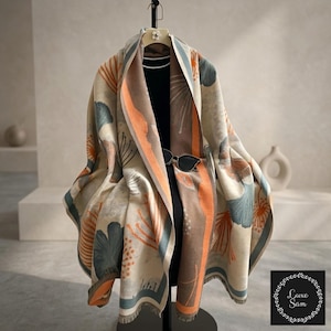 May include: A large, draped shawl featuring a cream base with a botanical pattern of teal and orange ginkgo-like leaves. The shawl has a prominent orange border and a fringed hem. A pair of black sunglasses rests on the fabric, which is displayed on a black garment form.