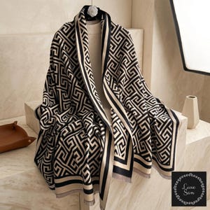 May include: A large, rectangular shawl featuring a repeating black and cream geometric Greek key pattern. The fabric has a thick, woven texture with a solid black border along the edges and a fringed hem. A small logo reads Luxe Sam.