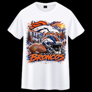 Puede incluir: Una camiseta blanca con un estampado gráfico del logotipo de los Denver Broncos, un balón de fútbol americano y un casco. El diseño incluye un paisaje montañoso, el horizonte de una ciudad y pinos con salpicaduras de pintura naranja y azul.