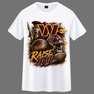 May include: A white short-sleeve t-shirt featuring a graphic print of a Washington Commanders helmet, a shield, a spear, and a football. The design includes the team logo, the text "WASHINGTON COMMANDERS" and "RAISE HAIL" in gold and burgundy.