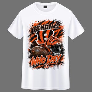 Puede incluir: Una camiseta blanca de manga corta con un estampado gráfico de un casco de fútbol americano, un balón y el texto BENGALS y WHO DEY en naranja y negro. El diseño incluye patrones de rayas de tigre y efectos de salpicaduras de pintura.