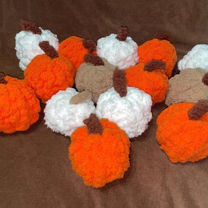 May include: A collection of handmade plush pumpkins crafted from soft, textured yarn. The pumpkins are available in vibrant orange, crisp white, and muted tan colors, each featuring a small, dark brown stem attached to the top of the round body.