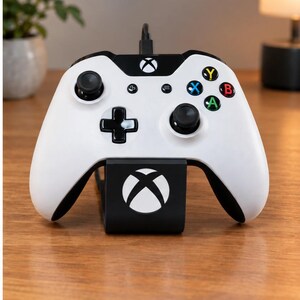May include: A white and black video game controller resting on a black charging stand. The controller features a central Xbox logo, two black analogue sticks, a directional pad, and four coloured action buttons labelled X, Y, B, and A.
