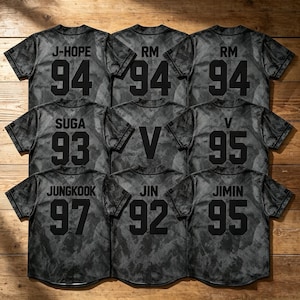 Op de afbeelding: Een collectie van negen honkbalshirts met korte mouwen in een donkergrijs camouflagepatroon. Elk shirt heeft een naam en een tweecijferig nummer in het zwart op de achterkant, waaronder J-HOPE 94, RM 94, SUGA 93, V, V 95, JUNGKOOK 97, JIN 92 en JIMIN 95.
