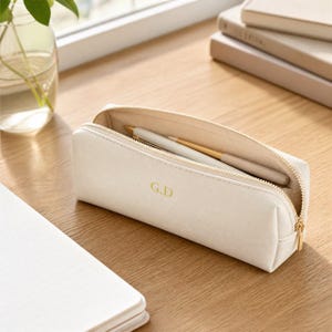 May include: A cream-colored rectangular pencil case with a gold zipper closure. The exterior features the gold-embossed initials G.D. The case is open, revealing a single light-colored pen resting inside the main compartment on a light interior lining.