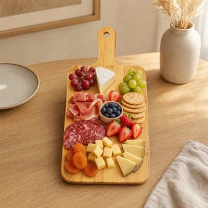 Puede incluir: Una tabla de servir rectangular de bambú con asa, cubierta con una selección de embutidos, uvas rojas y verdes, rodajas de salami, prosciutto, brie, cubos de queso cheddar, fresas, arándanos, albaricoques secos y galletas saladas redondas.
