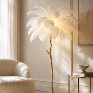 Op de afbeelding: Een vloerlamp met een goudkleurige voet in de vorm van een getextureerde boomtak met een driepotige basis. De bovenkant van de lamp is versierd met talloze witte struisvogelveren die een warm, zacht licht vanuit het midden verspreiden.