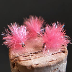 May include: Three bright pink fishing flies with iridescent fibers attached to small metal hooks. The flies feature a fluffy, textured appearance with fine, shimmering strands that catch the light, creating a vibrant and eye-catching display.