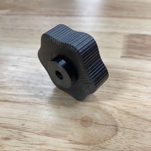 May include: A black, multi-lobed control knob with a textured, ribbed outer edge and a central circular hub featuring a splined, star-shaped internal opening. The object has a layered, 3D-printed appearance with visible horizontal lines.