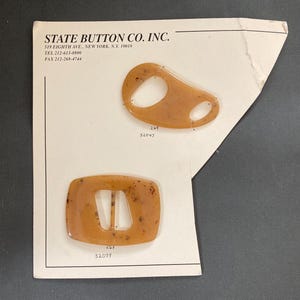 May include: Two vintage brown plastic buckles on a white card from State Button Co. Inc. One buckle is a rounded rectangle with a center opening, the other is an oval shape with a hole. The card has the company's address and phone number printed on it.