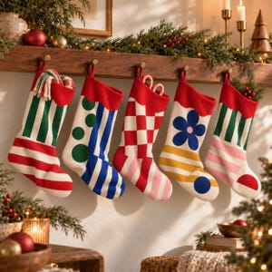 Op de afbeelding: Vijf gebreide kerstsokken hangen op een rij aan een houten schoorsteenmantel. De sokken hebben verschillende patronen, waaronder groen-witte strepen, blauwe stippen en strepen, een rood-wit dambordpatroon, een blauwe bloem en groene strepen.