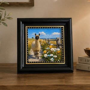 May include: A square framed painting featuring a cream-colored llama standing on a dirt path in a field of yellow and white wildflowers under a bright blue sky. The artwork is encased in a thick black frame with a gold inner border and studs.