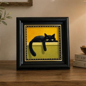 Könnte beinhalten: Ein quadratisch gerahmtes Gemälde, das eine schwarze Katze zeigt, die auf einem Sims vor einem gelben Hintergrund liegt. Die Katze hat gelbe Augen und ihr Schwanz hängt über die Kante. Der Rahmen ist schwarz mit einem goldenen Innenrand.