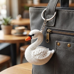 May include: A white crocheted swan keychain with an orange beak and black eye detail, attached to a silver metal split ring and chain, hanging from the zipper pull of a dark gray canvas tote bag featuring a front pocket and brass hardware.
