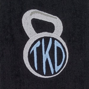 May include: Black fitness towel with a silver metal loop for hanging. The towel features an embroidered kettlebell design with the letters "TKD" in light blue and gray.