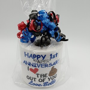 May include: A novelty gift of a roll of toilet paper wrapped in cellophane and topped with red, blue, and black curling ribbon. The roll is decorated with the text "Happy 1st Anniversary" and a heart and poop emoji.