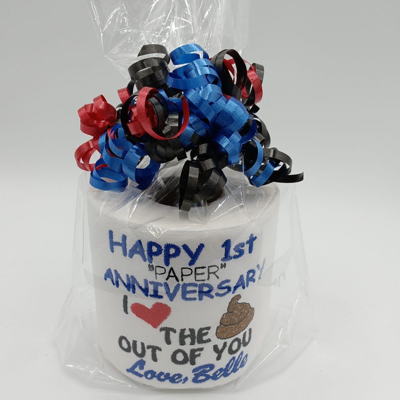 May include: A novelty gift of a roll of toilet paper wrapped in cellophane and topped with red, blue, and black curling ribbon. The roll is decorated with the text "Happy 1st Anniversary" and a heart and poop emoji.