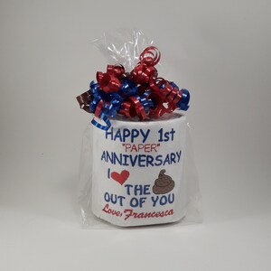 May include: A novelty gift of a toilet paper roll wrapped in cellophane, topped with red, white, and blue ribbons. The roll is embroidered with the words "Happy 1st 'Paper' Anniversary" and "I love the out of you".