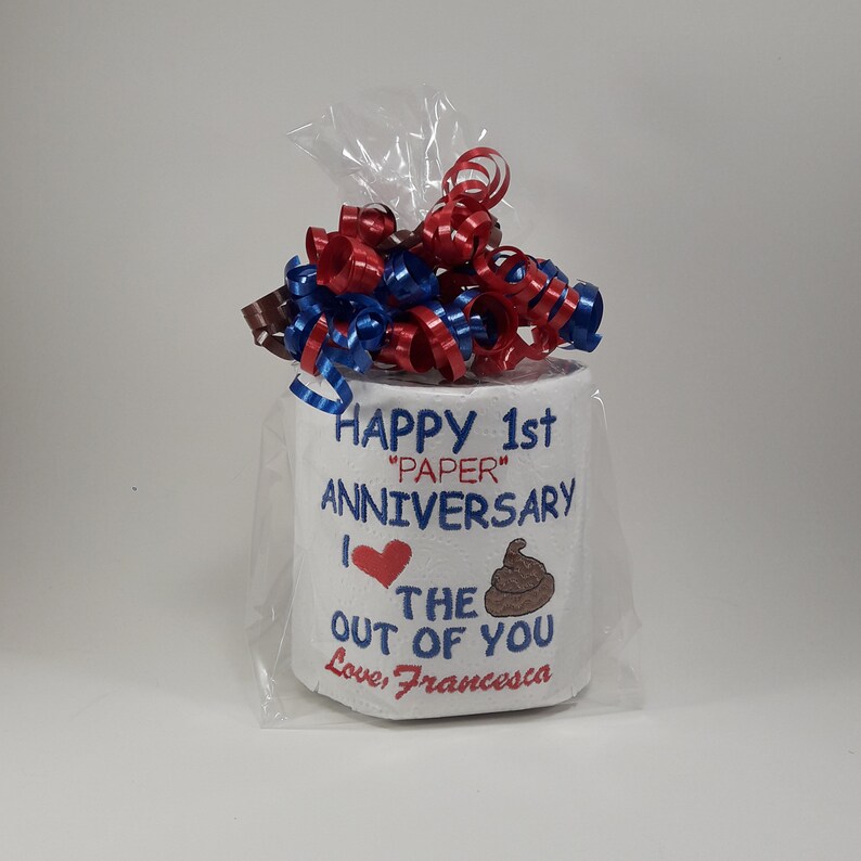 May include: A novelty gift of a toilet paper roll wrapped in cellophane, topped with red, white, and blue ribbons. The roll is embroidered with the words "Happy 1st 'Paper' Anniversary" and "I love the out of you".