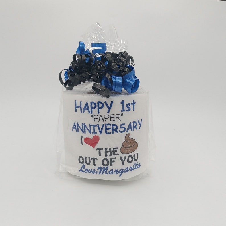 May include: A roll of white toilet paper wrapped in cellophane, decorated with blue and black ribbon. The roll is inscribed with the words "HAPPY 1st 'PAPER' ANNIVERSARY" and a heart and poop emoji. The message continues "I ❤️ THE OUT OF YOU Love, Margarita."