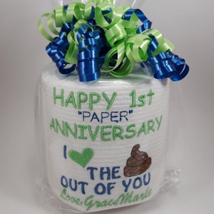May include: A roll of white toilet paper is wrapped in clear cellophane and topped with blue and green ribbon. The roll is embroidered with the words "HAPPY 1st 'PAPER' ANNIVERSARY" and a heart and poop emoji. The text "I the out of you" is also embroidered.