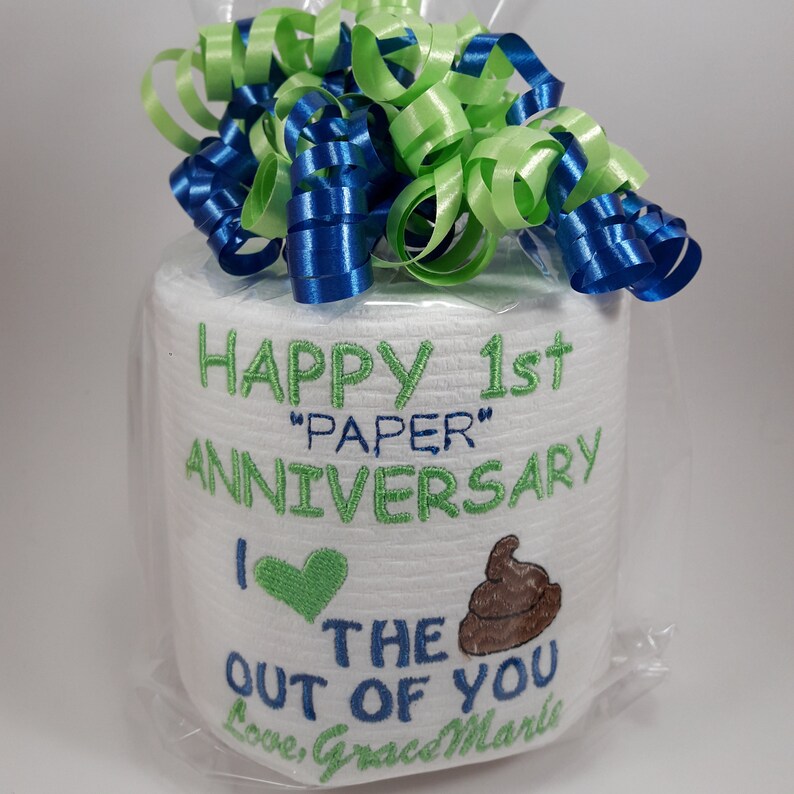 May include: A roll of white toilet paper is wrapped in clear cellophane and topped with blue and green ribbon. The roll is embroidered with the words "HAPPY 1st 'PAPER' ANNIVERSARY" and a heart and poop emoji. The text "I the out of you" is also embroidered.