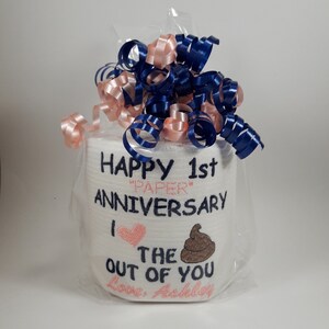 May include: A roll of white toilet paper is wrapped in clear cellophane and topped with blue and pink ribbons. The roll is embroidered with the words "HAPPY 1st ANNIVERSARY" and "I the out of you Love, Ashley."