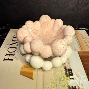 May include: A decorative bowl featuring a top section with a marbled peach and white finish and a bottom section composed of solid off-white spheres. The bowl has a rounded, bubble-like texture throughout its structure, creating a distinct layered appearance.
