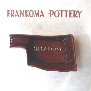 May include: A brown glazed ceramic dish shaped like the state of Oklahoma with the word OKLAHOMA embossed in the center. Above the dish, the text FRANKOMA POTTERY is printed in a brown sans-serif font on a plain white background.