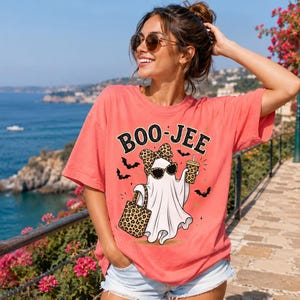 May include: A coral-colored t-shirt featuring a graphic of a white ghost wearing leopard-print sunglasses and a matching bow, holding a leopard-print tote bag and a drink. The text BOO-JEE is printed above the ghost, surrounded by small bats.