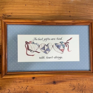 Peut inclure: Un cadre en bois rectangulaire contenant une impression sur fond bleu clair à pois blancs. L'image montre trois cœurs au motif patchwork bleu, blanc et rouge, reliés par des rubans rouges, avec le texte : The best gifts are tied with heart-strings.
