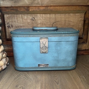 May include: A rectangular light blue train case with a top handle, featuring a central silver-toned metal latch and a small rectangular plaque at the bottom center with the text Fashion Flight embossed in a script font on the front surface.