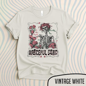 May include: Vintage white t-shirt featuring a skeleton with a floral crown and roses, with the words "Grateful Dead" in a decorative font. The shirt has rolled-up sleeves and a crew neck.