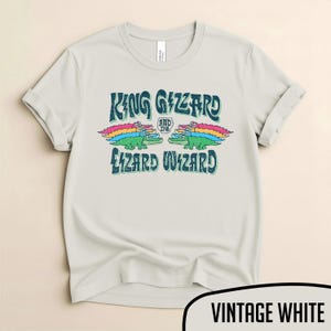 May include: Vintage white t-shirt featuring the text "KING GIZZARD AND THE LIZARD WIZARD" in a blue, retro font. The design includes two green alligators with rainbow wings. A casual, unisex tee.
