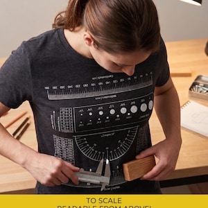May include: A dark gray t-shirt with a ruler and measurement diagrams printed on it. The text "TO SCALE READABLE FROM ABOVE!" is at the bottom. A person is using a caliper to measure a wooden block.