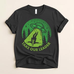 May include: Black t-shirt with a green graphic of a UFO beaming up a person. The design includes green trees and the text "TAKE OUR LEADER" in green, set within a green circle.