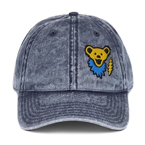 May include: A denim blue baseball cap with a distressed finish. The cap features an embroidered design of a yellow bear with a blue collar and a yellow lightning bolt. The cap has a curved brim and an adjustable strap.