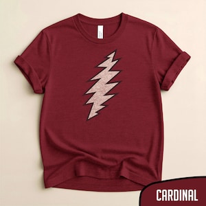 May include: A cardinal red t-shirt featuring a stylized lightning bolt design in light brown with a black outline. The short-sleeved shirt has rolled-up sleeves and a crew neck. The word "CARDINAL" is in a white font on a dark red background.