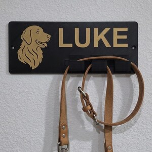 May include: A rectangular black wall-mounted leash holder featuring a gold-colored silhouette of a dog head and the name LUKE in gold lettering. The holder has three black hooks, with a tan leather leash and collar hanging from the hooks.