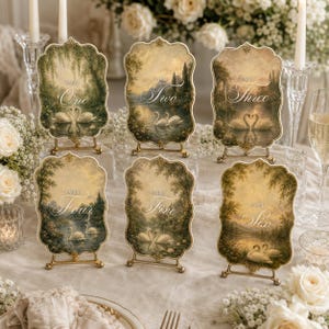 Pode incluir: Seis placas decorativas de mesa com bordas recortadas, cada uma apresentando uma pintura de paisagem única de cisnes em um lago. As placas estão rotuladas de Table One a Table Six em escrita branca e exibidas em pequenos suportes dourados.