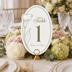 Może przedstawiać: Owalna karta z numerem stołu ozdobiona ilustracją zielonej kalii, napisem "Table 1" oraz imionami "Mariana & Daniel". Karta jest umieszczona pionowo w małym, okrągłym, złotym metalowym stojaku na białym tle.