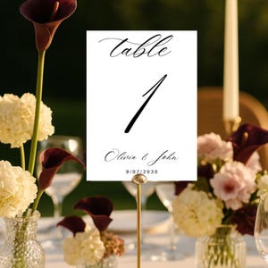 Pode incluir: Um cartão de número de mesa retangular branco com a palavra table em letra cursiva preta, o número 1 em fonte preta grande, e os nomes Olivia & John com a data 9/07/2030 impressos em preto na parte inferior, num suporte de metal dourado.