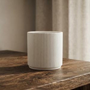 May include: A cylindrical white ceramic planter featuring a vertical ribbed texture along the exterior surface. The planter has a flat base and an open top, showcasing a clean, uniform design with a matte finish throughout the entire structure.
