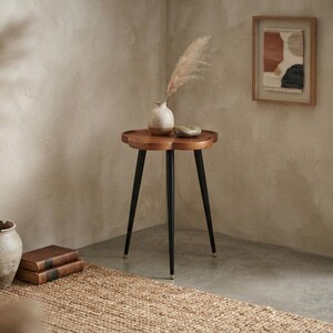 May include: A small, round, wooden side table with a scalloped edge and three slender, black, tapered legs. The table features a smooth, polished brown wood top and is supported by black metal legs with small, gold-toned metallic feet.