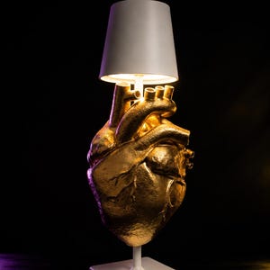Könnte beinhalten: Eine Tischleuchte mit einem goldfarbenen anatomischen Herz als Lampenfuß. Die Leuchte verfügt über einen weißen quadratischen Sockel, einen weißen vertikalen Ständer und einen weißen konischen Lampenschirm, der warmes Licht ausstrahlt.