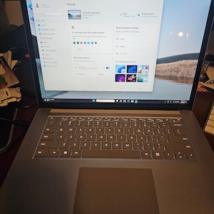 Puede incluir: Un portátil plateado con teclado retroiluminado y un panel táctil grande, que muestra el menú de configuración de Windows en su pantalla. La pantalla muestra la pestaña Inicio con información del dispositivo y opciones de personalización.