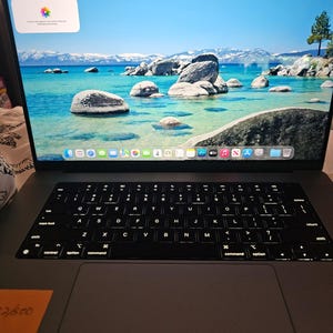 Puede incluir: Un portátil gris espacial con teclado QWERTY retroiluminado y un trackpad grande. La pantalla muestra una interfaz de escritorio con un fondo de pantalla de un lago azul, una costa rocosa y montañas, junto con widgets y iconos del sistema.