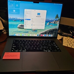 May include: A space grey MacBook Pro laptop with a black backlit keyboard and a large trackpad. The screen displays the macOS System Settings menu and an About window. A small orange sticky note with handwritten text is attached to the palm rest.
