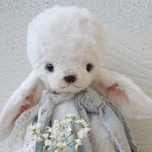 Könnte beinhalten: Ein weißer Plüschhase mit langen Ohren und einem blau-weißen Kleid. Der Hase hält einen Strauß weißer Blumen und sitzt auf einem weißen Hintergrund.